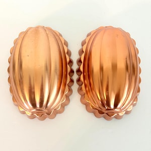May include: Two oval-shaped copper-colored baking molds with a metallic finish. Each mold features a series of vertical ridges running along the center and a scalloped, fluted edge around the perimeter, creating a distinct textured pattern.