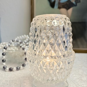 Puede incluir: Un portavelas de cristal transparente con un diseño de diamantes. El portavelas tiene una base y una tapa extraíble. Una cadena de perlas está en el fondo. El portavelas está encendido, proyectando un brillo cálido.