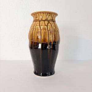 May include: A handcrafted ceramic vase with a two-tone glaze. The upper portion is a light brown with vertical drip patterns, transitioning to a dark brown base. The vase has a wide body and a slightly flared rim.