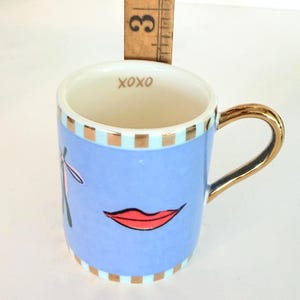 May include: A small, light blue ceramic mug with a gold handle and rim accents. The mug features a red lip graphic and the word "XOXO" inside. A wooden ruler is placed behind the mug, measuring approximately 7.6 cm.