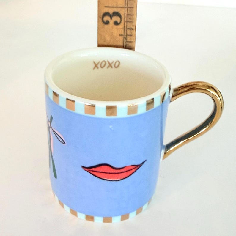 May include: A small, light blue ceramic mug with a gold handle and rim accents. The mug features a red lip graphic and the word "XOXO" inside. A wooden ruler is placed behind the mug, measuring approximately 7.6 cm.