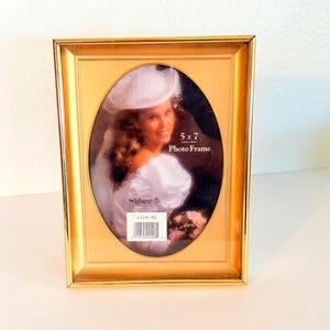 Peut inclure: Un cadre photo rectangulaire doré avec une zone d'affichage intérieure ovale. Le cadre contient un insert promotionnel montrant une personne en robe et chapeau blancs, avec le texte 5 x 7 Photo Frame et la marque Intercraft visibles.