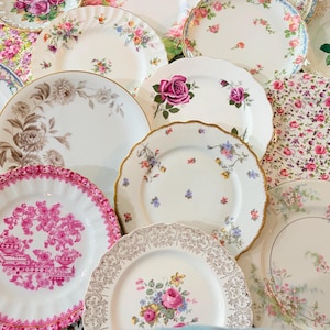 May include: A collection of assorted ceramic plates featuring various floral patterns, including pink roses, purple violets, and intricate gold filigree borders. The plates vary in size and shape, with some displaying scalloped edges and detailed botanical illustrations.