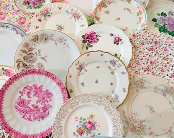 May include: A collection of assorted ceramic plates featuring various floral patterns, including pink roses, purple violets, and intricate gold filigree borders. The plates vary in size and shape, with some displaying scalloped edges and detailed botanical illustrations.