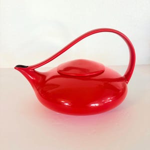 May include: A bright red teapot with a low, rounded body and a smooth, glossy finish. It features a wide, curved handle that extends from the top of the body to the spout, and a circular lid centered on the top of the vessel.