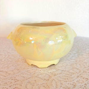 May include: A round, pale yellow ceramic bowl with an iridescent finish. The bowl has two small, pointed handles and sits on a low, scalloped base. The interior is a lighter shade of yellow.