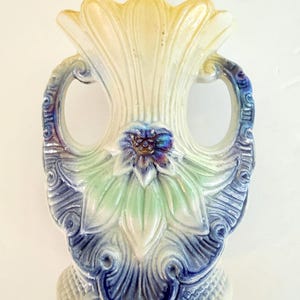 May include: A ceramic vase with a scalloped rim and two handles, featuring an iridescent finish. The body displays a molded floral design in shades of cream, pale green, and deep blue, with intricate scrollwork patterns at the base and handles.