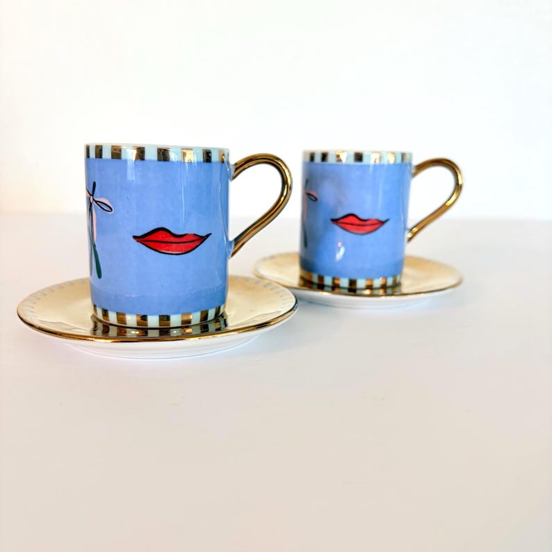 May include: Two blue ceramic coffee cups with gold handles and red lip designs, each on a matching saucer. The cups have a striped pattern at the top and bottom. The saucers and handles are gold.