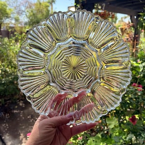 May include: A clear glass plate featuring a scalloped edge and a radial sunburst pattern radiating from the center. The design includes a series of embossed, fan-shaped panels around the perimeter and a textured, geometric starburst at the middle.