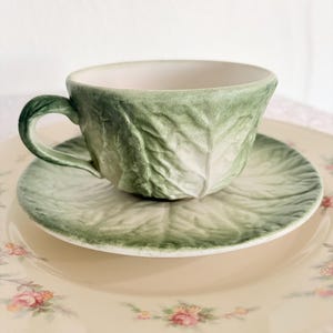 Puede incluir: Un juego de taza y platillo de cerámica verde, con forma y textura de hojas de col. La taza se apoya en un platillo a juego, colocado sobre un plato floral con rosas rosas y un borde dorado. Un juego de té único.