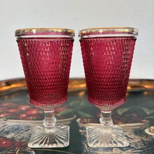 May include: Two ruby red vintage wine glasses with a diamond pattern and gold rim. The glasses have a square base and a clear stem. The glasses are displayed on a decorative tray.