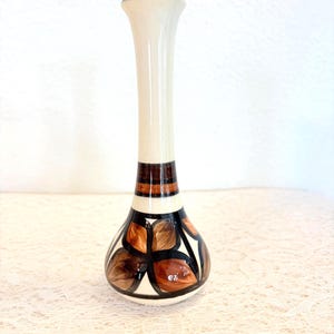Peut inclure: Un vase en céramique, élancé et de forme allongée, avec une partie supérieure crème et une base bulbeuse. La base présente un motif de feuilles marron et beige souligné de noir. Le vase a une bande marron près du haut et un bord marron foncé.