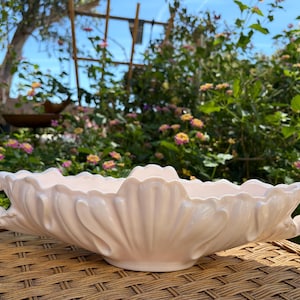 May include: A light pink, ceramic serving dish with a scalloped edge and two ornate handles. The dish has a shell-like design and sits on a woven surface, with a garden background.