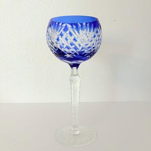 May include: A clear crystal wine glass with a cobalt blue rim. The bowl features a diamond-cut pattern, and the stem is clear. The glass sits on a clear, circular base. The glass is designed for serving wine.