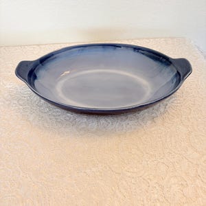 May include: A blue oval serving dish with two handles. The dish has a gradient effect, transitioning from a darker blue at the edges to a lighter blue in the center. The dish is on a cream-colored lace tablecloth.