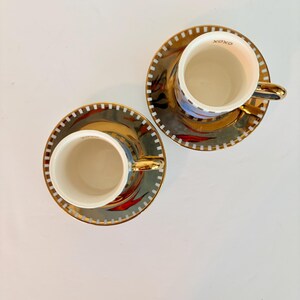 May include: Two matching teacups and saucers. The cups and saucers have a gold rim and handle. The saucers have a gold and black design with red accents. The cups have the word "XOXO" printed on the rim.