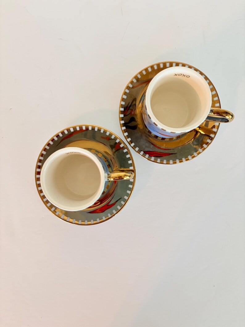 May include: Two matching teacups and saucers. The cups and saucers have a gold rim and handle. The saucers have a gold and black design with red accents. The cups have the word "XOXO" printed on the rim.