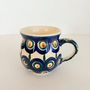 May include: A ceramic mug with a rounded body and a handle, featuring a hand-painted pattern of concentric circles in shades of dark blue, green, and light orange against a cream-coloured background. The mug has a smooth, glazed finish.