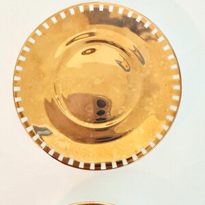 May include: Two gold-coloured plates with a shiny finish. Each plate has a white, rectangular pattern around the rim. The plates are round and appear to be made of metal. The plates are likely for decorative or serving purposes.