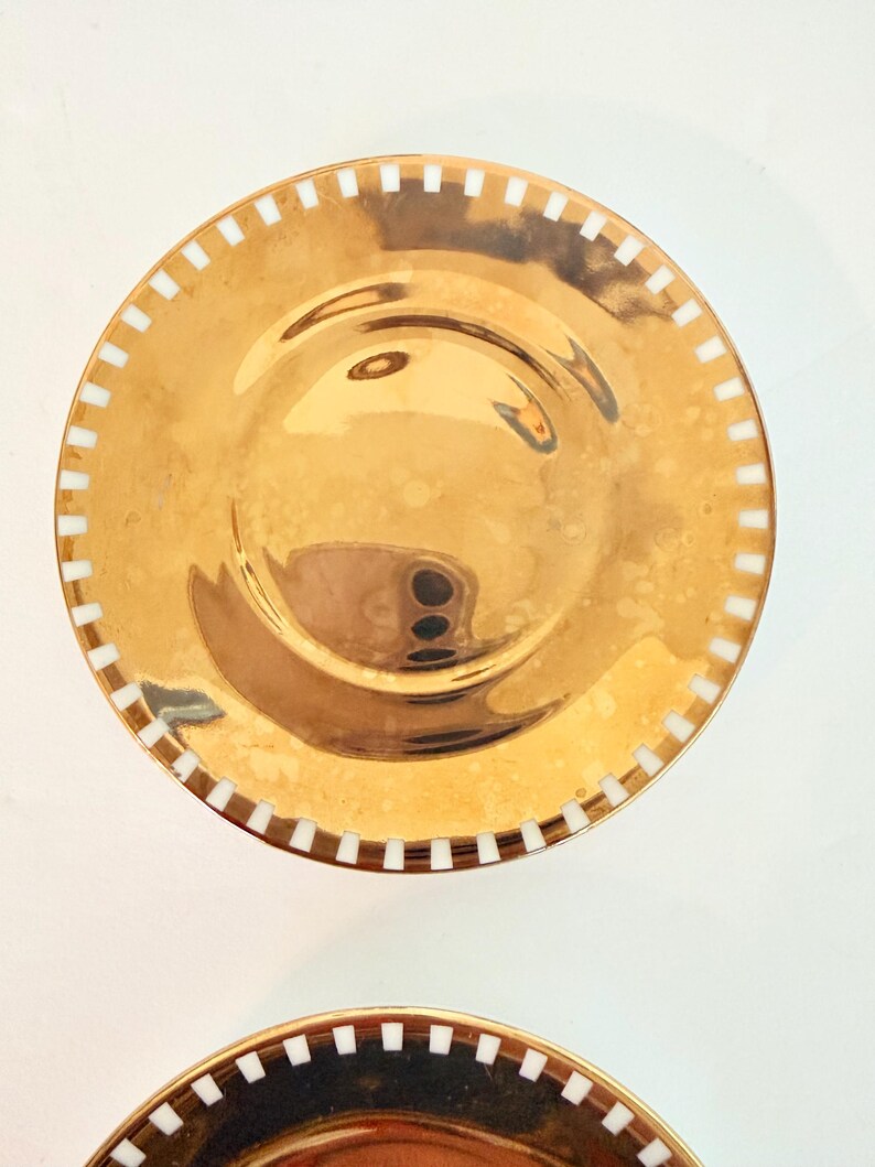 May include: Two gold-coloured plates with a shiny finish. Each plate has a white, rectangular pattern around the rim. The plates are round and appear to be made of metal. The plates are likely for decorative or serving purposes.