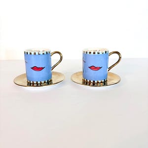 May include: A pair of blue espresso cups with gold handles and saucers. Each cup has a red lip design and black and white striped trim. The saucers have a gold metallic finish, creating a stylish coffee set.