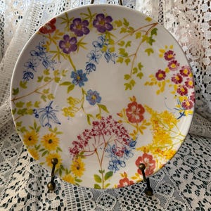 May include: White ceramic plate with a colorful floral design. The plate features a variety of flowers in shades of purple, red, yellow, and blue, with green leaves. The plate is displayed on a decorative metal stand.