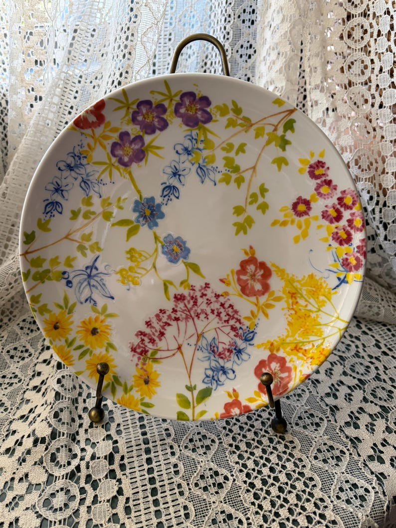 May include: White ceramic plate with a colorful floral design. The plate features a variety of flowers in shades of purple, red, yellow, and blue, with green leaves. The plate is displayed on a decorative metal stand.