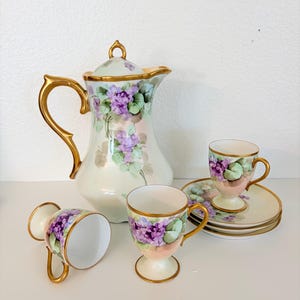 May include: A porcelain tea set featuring a tall pitcher with a lid, three cups, and three saucers. Each piece is decorated with hand-painted purple violet flowers and green leaves on a cream background, accented with gold trim on the handles and rims.