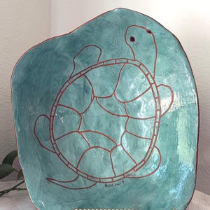 Könnte beinhalten: Handgefertigte, türkisfarbene Keramikschale mit Schildkrötendesign. Die Schildkrötenkontur ist rotbraun, und die Schale hat eine einzigartige, organische Form. Die Signatur des Künstlers, "kochut", ist auf der Schale sichtbar.