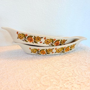 Puede incluir: Dos fuentes de servir oblongas de color blanquecino con un estampado floral en naranja, amarillo y verde. Las fuentes tienen un asa en un extremo y están apiladas. El diseño floral se repite en ambas fuentes. Cada fuente mide unos 25 cm de largo.