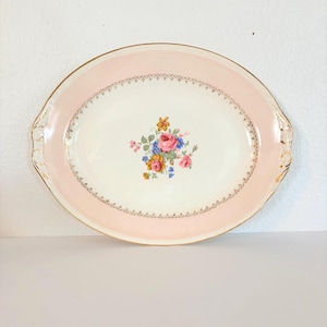 Könnte beinhalten: Ovale Servierplatte mit rosa Rand und einem Blumenstrauß in der Mitte. Die Platte hat goldfarbene Verzierungen und Griffe. Die Blumen sind rosa, blau und gelb. Die Platte ist wahrscheinlich aus Keramik.