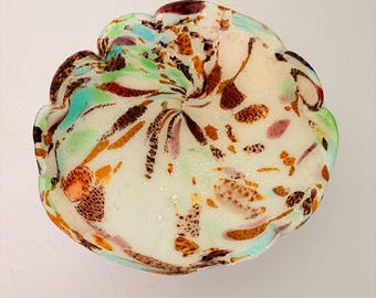 May include: A round glass bowl with a scalloped edge featuring a white base color. The surface is decorated with a speckled pattern of brown, orange, teal, and purple glass fragments arranged in a swirling design radiating from the center.