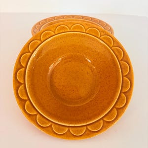 Op de afbeelding: Twee ronde keramische kommen in een warme mosterdgele kleur met een gespikkelde afwerking. Elke kom heeft een verhoogd schulprandpatroon rond de rand. De kommen zijn gestapeld, waarbij de ene kom direct voor de andere is geplaatst.