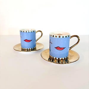 May include: Two blue espresso cups with gold handles and saucers. Each cup features a red lip design and a gold and black checkered rim. The saucers are gold. The cups have the text "XOXO" on the rim.