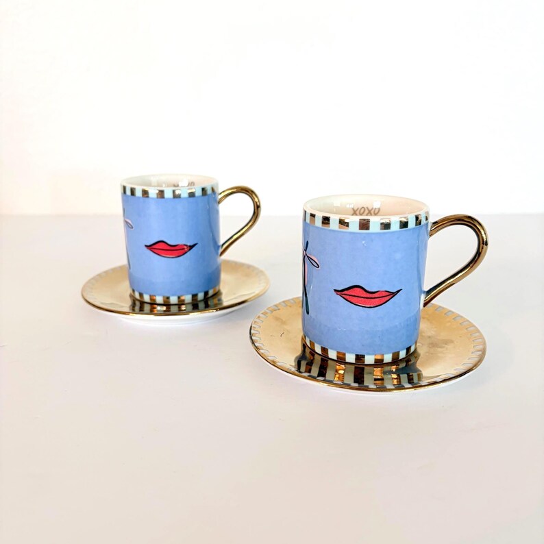 May include: Two blue espresso cups with gold handles and saucers. Each cup features a red lip design and a gold and black checkered rim. The saucers are gold. The cups have the text "XOXO" on the rim.