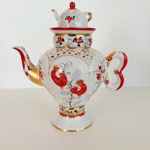 May include: A white porcelain teapot featuring a smaller teapot-shaped lid, decorated with red rooster illustrations, gold trim, and floral patterns. The vessel has a curved spout, a looped handle, and a pedestal base with intricate painted details.