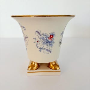 Puede incluir: Un jarrón de cerámica color crema con borde acampanado y acabado dorado. El cuerpo presenta un patrón floral repetitivo en azul y rojo. La base es cuadrada y se apoya en cuatro pies dorados, con una línea dorada que acentúa el borde.