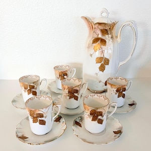 May include: A porcelain tea set consisting of a tall lidded pitcher and five matching cups with saucers. Each piece features a white base with a delicate gold leaf pattern and scalloped edges. The pitcher has a curved handle and a pointed spout.
