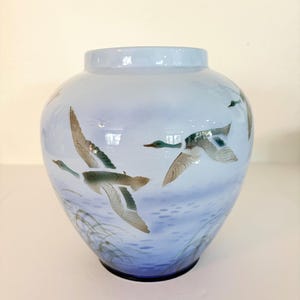 May include: A round glass vase with a light blue gradient background featuring a hand-painted scene of ducks in flight over water with reeds. The vase has a smooth, glossy finish and a wide opening at the top, showcasing detailed avian illustrations.