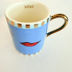 May include: A ceramic mug with a light blue exterior, featuring a red lip design and gold accents on the handle and rim. The interior is cream-coloured, with the word "XOXO" in gold. The mug has a capacity of approximately 300 ml.