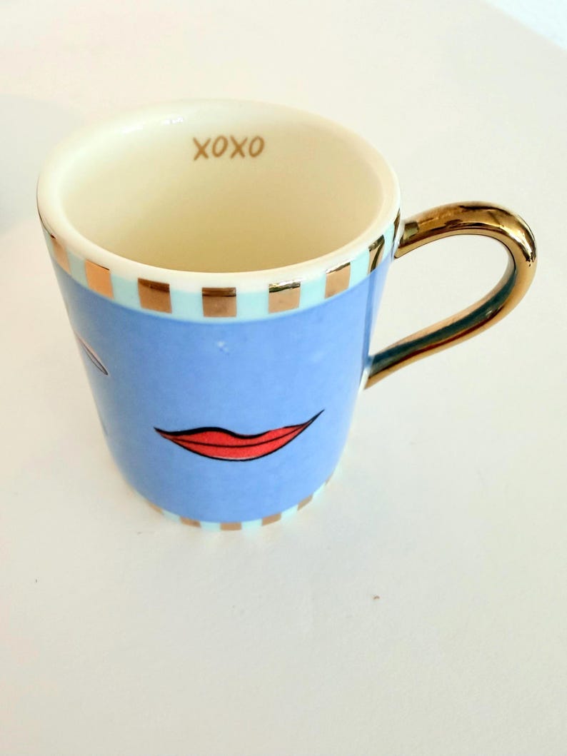May include: A ceramic mug with a light blue exterior, featuring a red lip design and gold accents on the handle and rim. The interior is cream-coloured, with the word "XOXO" in gold. The mug has a capacity of approximately 300 ml.
