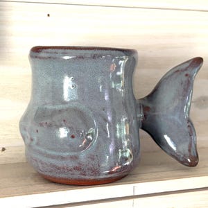 May include: A handmade ceramic mug in the shape of a fish. The mug is a muted blue-gray color with a fish tail handle. The rim and base of the mug are a reddish-brown color. The mug is sitting on a wooden shelf.