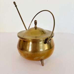 May include: A polished brass sugar bowl with a rounded body, three small feet, and a matching lid. The bowl features a thin, arched metal handle attached to the sides and includes a long, slender brass spoon resting inside the container.