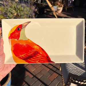 Puede incluir: Bandeja rectangular de cerámica de color crema. Presenta una ilustración de un pájaro vibrante en tonos rojos, naranjas y amarillos. El diseño del pájaro está en el lado izquierdo, mientras que el lado derecho permanece en blanco.