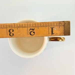 May include: A small, off-white teacup with a gold handle. A wooden ruler is placed across the cup's opening, measuring approximately 5 cm. The cup has a delicate, iridescent sheen.