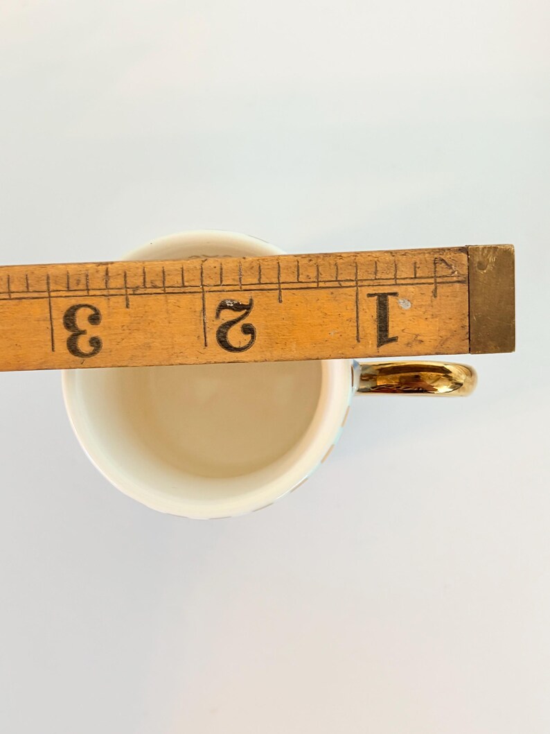 May include: A small, off-white teacup with a gold handle. A wooden ruler is placed across the cup's opening, measuring approximately 5 cm. The cup has a delicate, iridescent sheen.