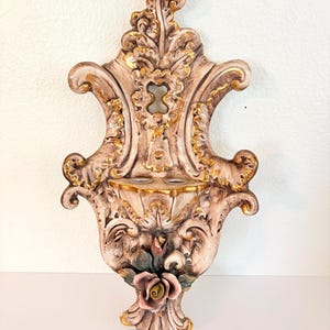 May include: A wall-mounted ceramic holy water font featuring an ornate, scroll-like design in shades of beige and brown with gold-painted accents. The piece includes a central basin and a sculpted pink rose with green leaves at the bottom.