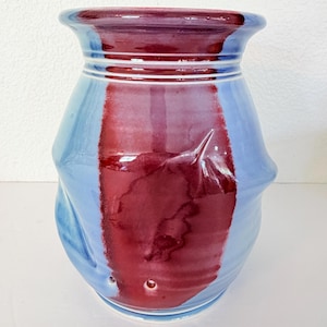 May include: A ceramic vase with a glossy finish, featuring a light blue base color and a wide, vertical stripe of deep maroon glaze running down the center. The vase has a flared rim, a slightly indented neck, and a rounded, textured body.