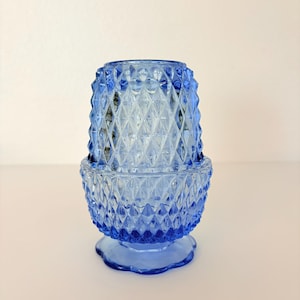 Puede incluir: Un portavelas de cristal azul con un diseño de diamantes. El portavelas consta de una base y una pieza superior, ambas con un diseño geométrico texturizado. La base tiene un borde festoneado.