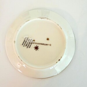 May include: Off-white Anthropologie plate with a gold rim. The base of the plate features a starburst design and the text "by ANTHROPOLOGIE". The plate has a slightly distressed appearance.
